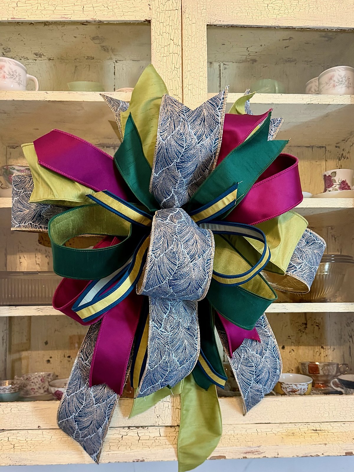 Handmade bow in magenta, forest green, chartreuse, and navy leaf-print ribbon, hanging against a cream crackle-painted china cabinet
