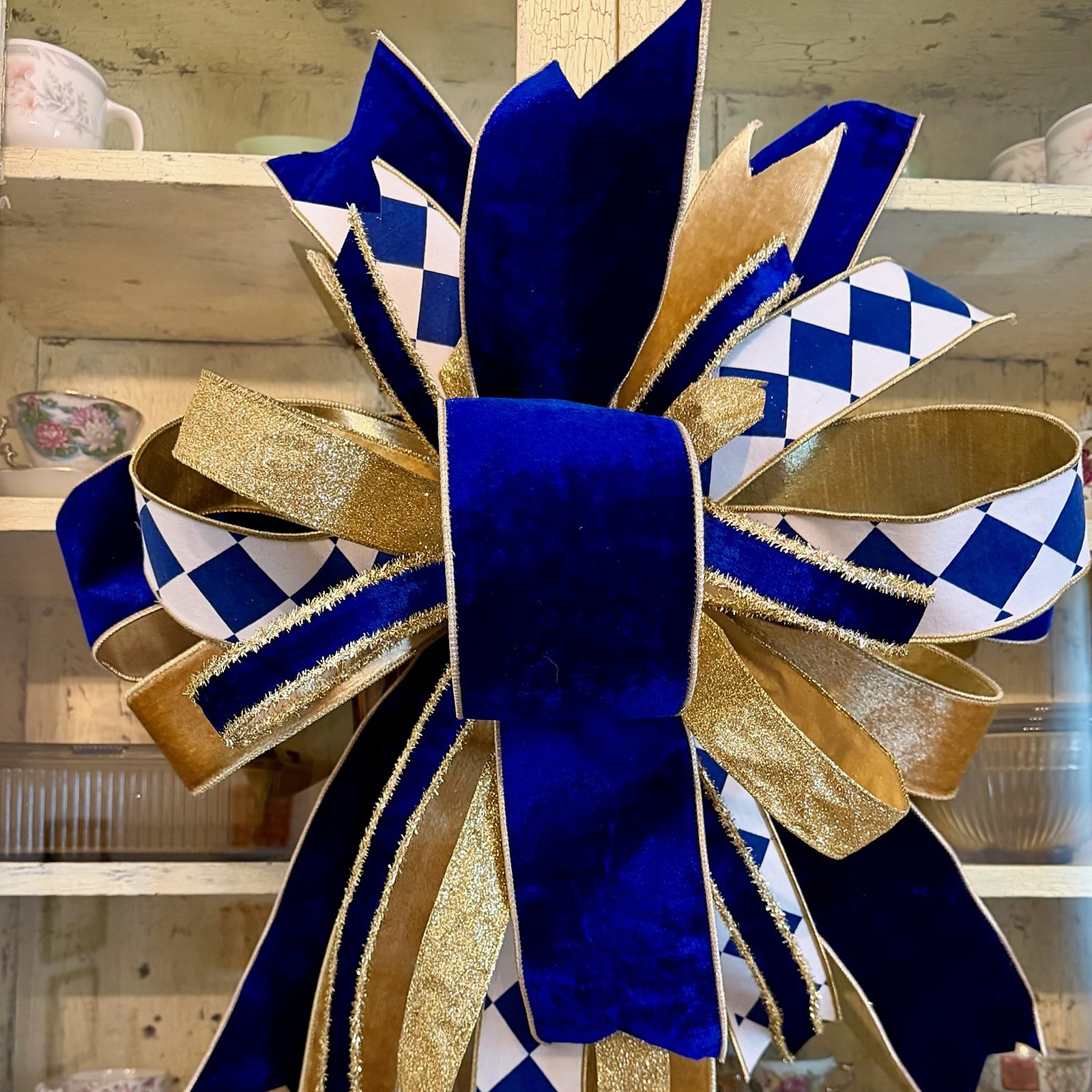 Handmade bow in royal blue velvet, gold glitter, and blue-and-white harlequin ribbon, hanging against a cream crackle-painted china cabinet